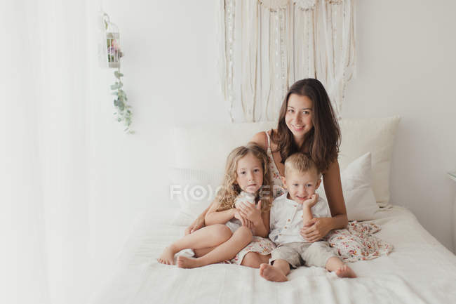 Позитивна молода брюнетка позує на ліжку як обіймає маленьких дітей чоловічої та жіночої статі в стильній спальні — стокове фото