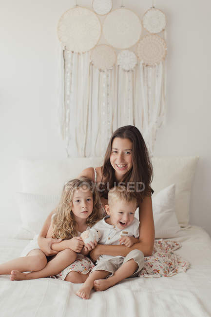 Позитивная юная брюнетка, позирующая на кровати, как обнимающая маленьких мальчиков и девочек в стильной спальне — стоковое фото