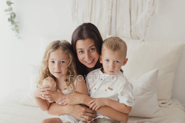 Portrait of young brunette sitting on bed and hugging little male and female children while looking in camera — Stock Photo