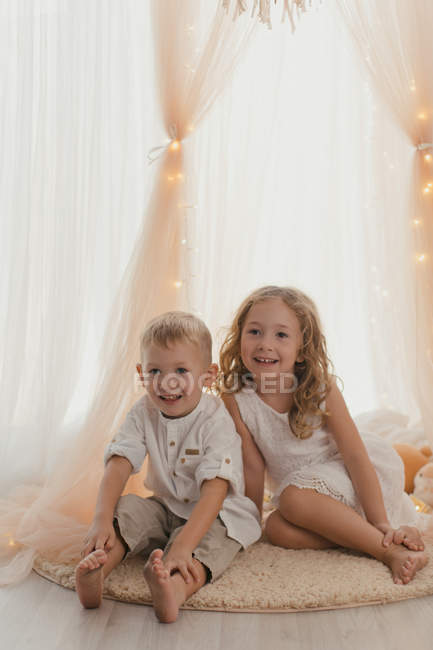 Bambina in abito bianco seduta su tappeto da maschio allegro bambino sorridente in macchina fotografica in camera elegante — Foto stock