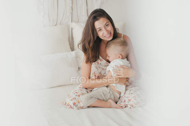 Удовлетворенная брюнетка в белом халате развлекается со счастливым мальчиком-малышом, обнимаясь на кровати — стоковое фото