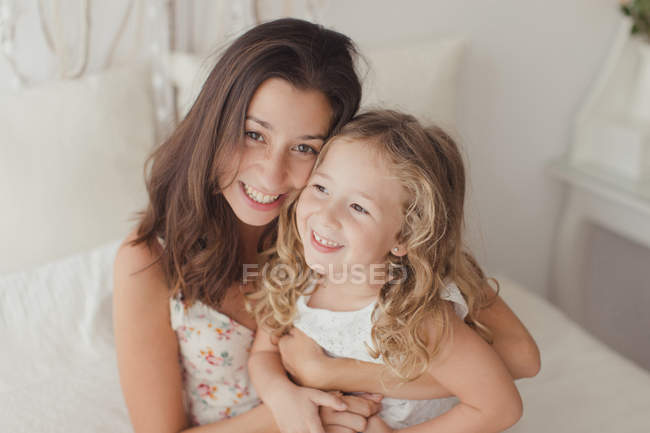 Glückliche brünette Mutter, die Spaß mit der blonden süßen Tochter hat, während sie sich im Bett umarmt — Stockfoto