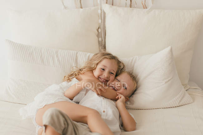 Little girl in white dress embracing male cheerful toddler brother on bed in stylish room — Stock Photo