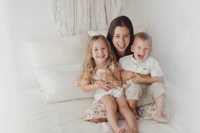 Positive young brunette sitting on bed and hugging little male and female children while looking in camera at stylish bedroom — Stock Photo