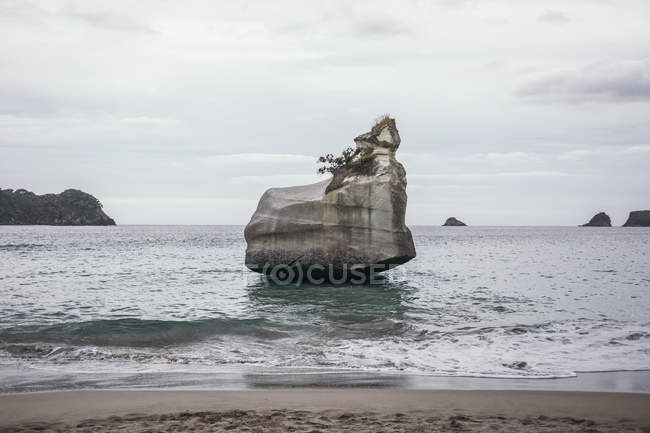 Gran roca entre el mar en el lado tropical solitario del océano en el tiempo nublado en la península de Coromandel en Nueva Zelanda - foto de stock