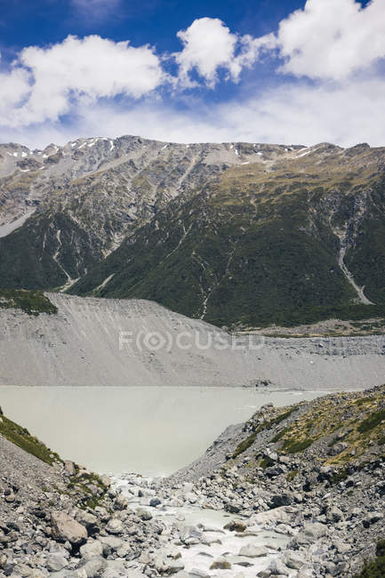 Мощные заросшие скалы возле маленького озера и большая снежная гора Кук с голубым горизонтом в Новой Зеландии — стоковое фото
