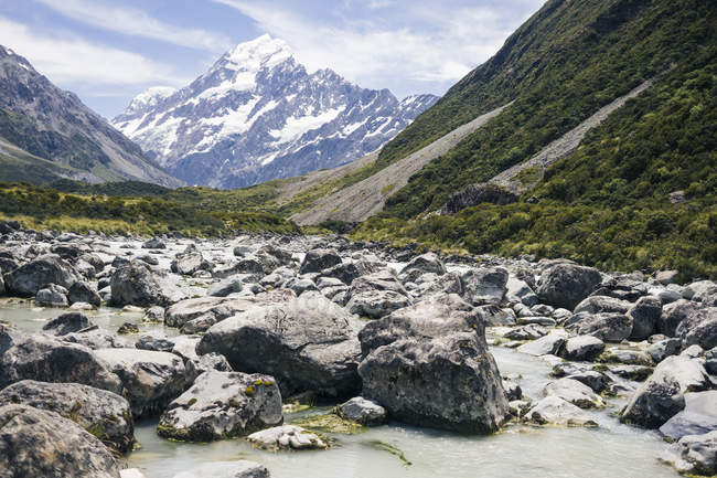 Río rocoso entre acantilados verdes con montaña Cook y cielo en Nueva Zelanda - foto de stock