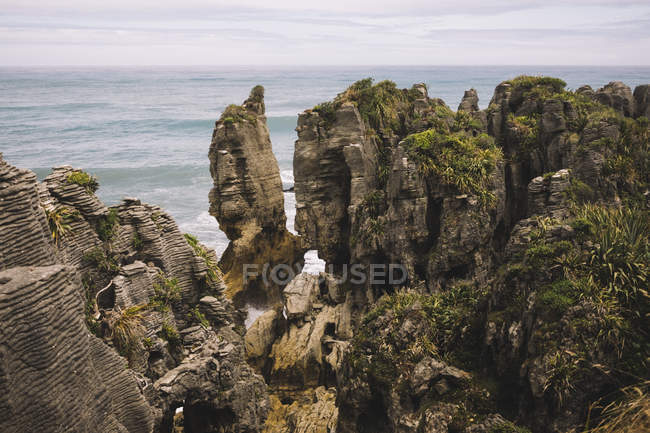 Desde arriba de rocoso cubierto de plantas a orillas del mar con olas y cielo nublado en Pancake Rocks en Nueva Zelanda - foto de stock