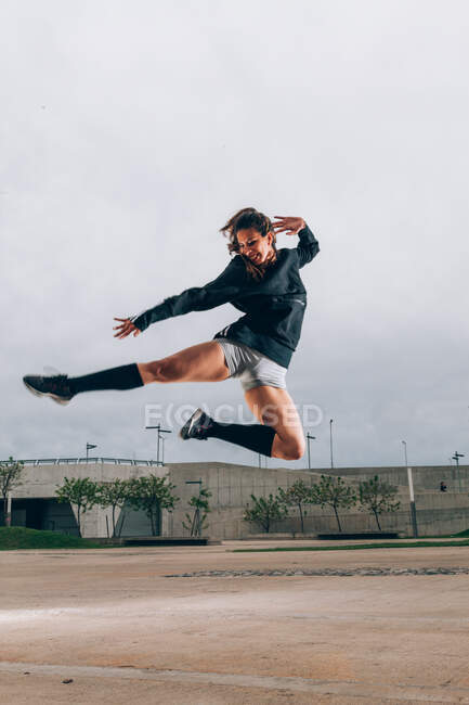 Joyful dançarina ajuste senhora em sportswear e tênis esticando as pernas e mão com o braço atrás da cabeça enquanto saltando ao ar livre campo de esportes — Fotografia de Stock