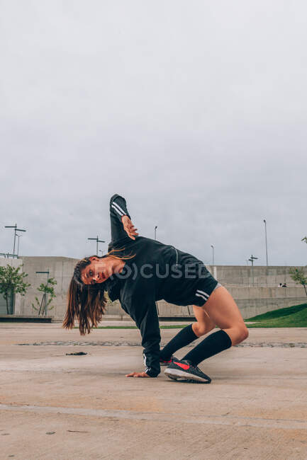 Концентрированная танцовщица хип-хопа в спортивной одежде, стоящая в позе моста на одной руке, глядя в камеру на пустую городскую улицу — стоковое фото
