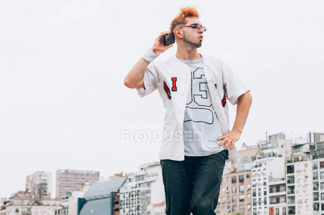 From below of stylish male hipster in trendy eyeglasses and casual clothing standing with arm akimbo while talking on mobile phone and looking away with sky and buildings on background — Stock Photo