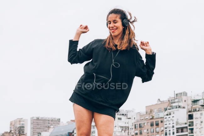 From below of joyful young female student in black hoodie standing with raised hands while listening music in headphones with buildings and sky on background — Stock Photo