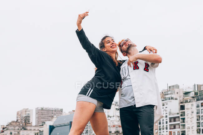 From below of young satisfied male and female in casual clothing standing and taking selfie on mobile phone with city and sky on background — Stock Photo