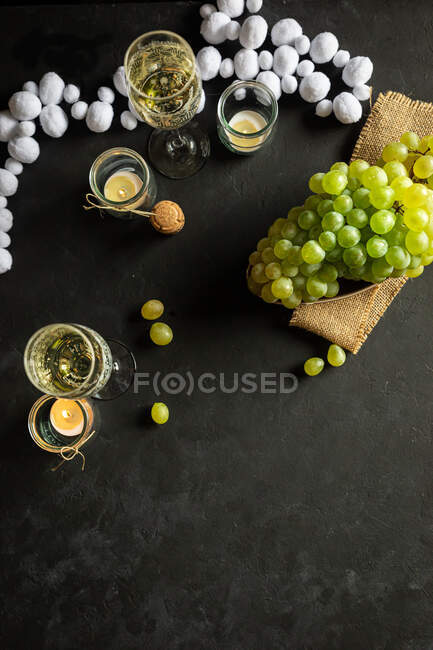 Празднование Нового года с бокалами вина с шампанским и зеленым виноградом изнасилования на столе украшены свечами чая и белой гирляндой на черном фоне — стоковое фото