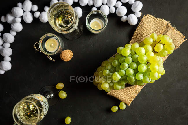 Celebración de Año Nuevo con copas de vino con champán y uvas verdes de colza en la mesa decoradas con velas de té y guirnalda blanca sobre fondo negro - foto de stock