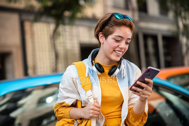 Adolescente moderno com fones de ouvido e bolsa de surf telefone celular enquanto sentado contra carros coloridos e edifícios modernos — Fotografia de Stock