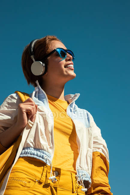 Low angle of bright millennial female in sunglasses and vivid white and yellow outfit wearing headphones against blue sky — Stock Photo