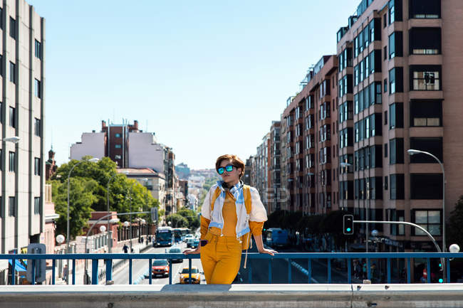 Trendy female with backpack standing on bridge against buildings under clear blue sky — Stock Photo