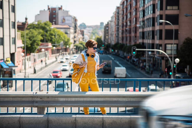 Femme en vêtements blancs et jaunes avec sac à dos debout sur le pont et regardant le téléphone portable contre la rue et les bâtiments — Photo de stock