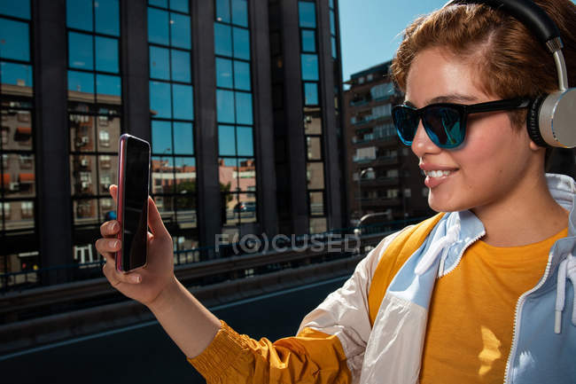 Улыбающийся модный подросток в солнечных очках в наушниках делает селфи со смартфоном против размытого современного здания — стоковое фото