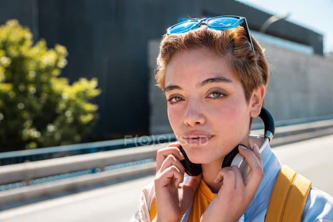 Großartige stylische Millennial-Frau mit Kopfhörern und Sonnenbrille, die in die Kamera gegen die verschwommene Straße schaut — Stockfoto