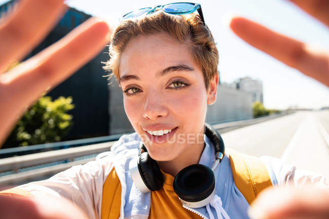 Magnífica mulher milenar elegante com fones de ouvido e óculos de sol olhando na câmera e mostrando as mãos contra a estrada — Fotografia de Stock