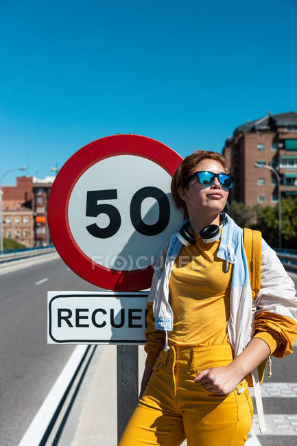Stylish happy pensive female in sunglasses and yellow clothes contemplating while standing beside restrictive traffic sign — Stock Photo