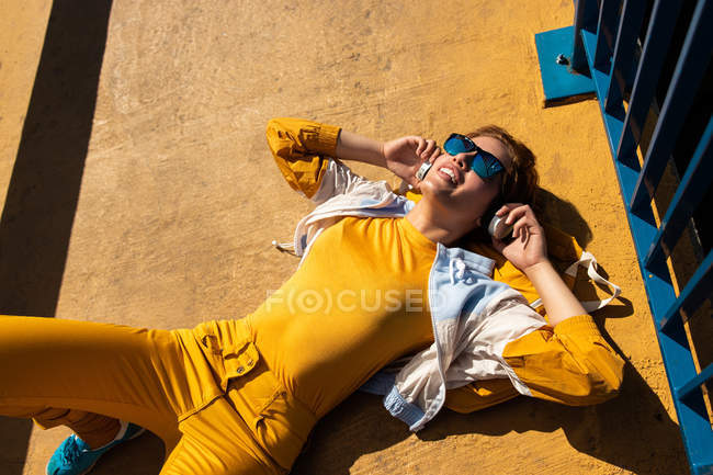 Сверху холодный подросток в солнечных очках слушает музыку в наушниках, лежа на ярком тротуаре с голубым забором — стоковое фото