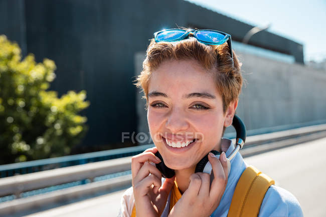 Magnificent stylish millennial woman with headphones and sunglasses looking in camera against blurred road — Stock Photo