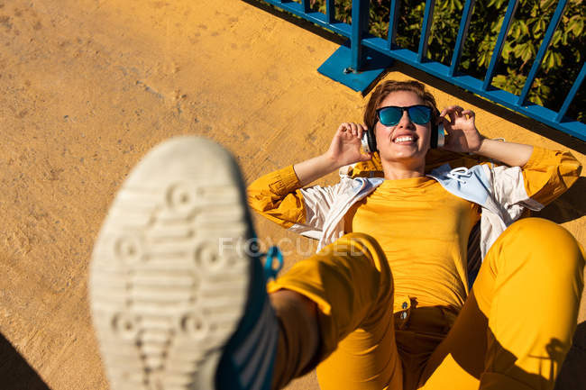 Сверху холодный подросток в солнечных очках слушает музыку в наушниках, лежа на ярком тротуаре с голубым забором — стоковое фото