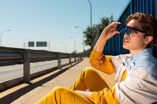 Adolescente escalofriante de moda en gafas de sol y ropa de color amarillo brillante sentado en el pavimento vívido con valla azul - foto de stock