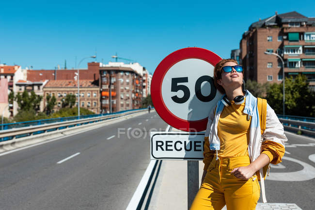 Стильна щаслива пенсіонерка в сонцезахисних окулярах і жовтому одязі, що розмірковує, стоячи поруч з обмежувальним дорожнім знаком — стокове фото