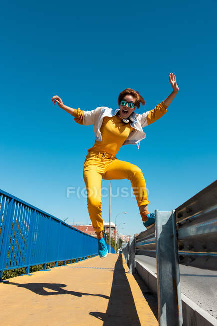 D'en bas courageux adolescent joyeux dans des lunettes de soleil sautant avec la bouche ouverte et les bras levés dans la rue — Photo de stock