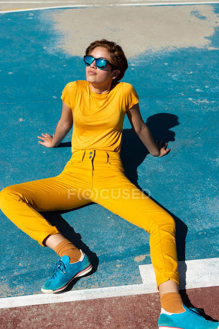 High angle of frowning cool female teenager in yellow clothes with sunglasses sitting on blue sports ground — Stock Photo