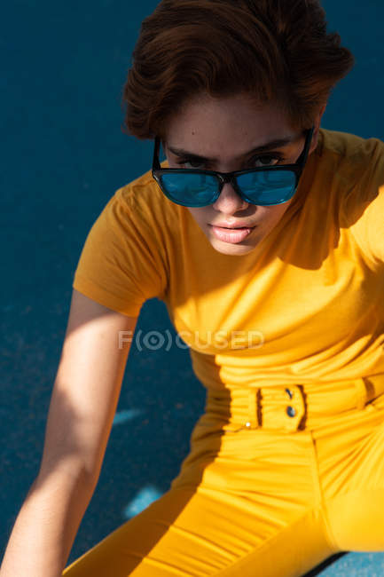 High angle of frowning cool female teenager in yellow clothes looking in camera with sunglasses — Stock Photo