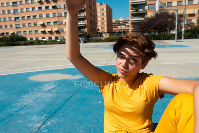 Alto ángulo de fruncir el ceño adolescente fresco en ropa amarilla que cubre la cara de la luz del sol con la mano mientras está sentado en el campo de deportes azul - foto de stock
