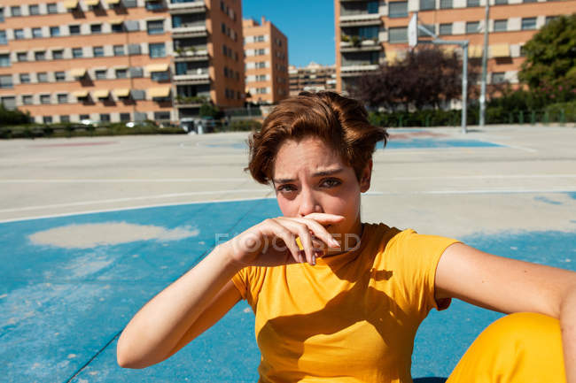 High angle of frowning female in yellow clothes looking in camera and covering mouth while sitting on blue sports ground — Stock Photo