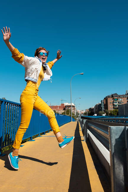 Da sotto adolescente coraggioso allegro in occhiali da sole che saltano con bocca aperta e braccia alzate su strada — Foto stock