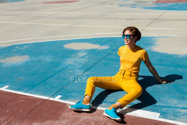 Angle élevé d'adolescente fraîche en vêtements jaunes avec des lunettes de soleil assis sur un terrain de sport bleu à la lumière du soleil — Photo de stock