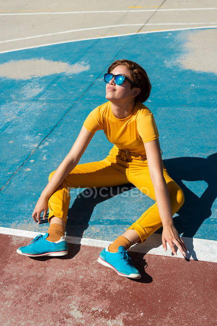 Alto angolo di cool adolescente in abiti gialli con occhiali da sole seduti su un terreno sportivo blu alla luce del sole — Foto stock