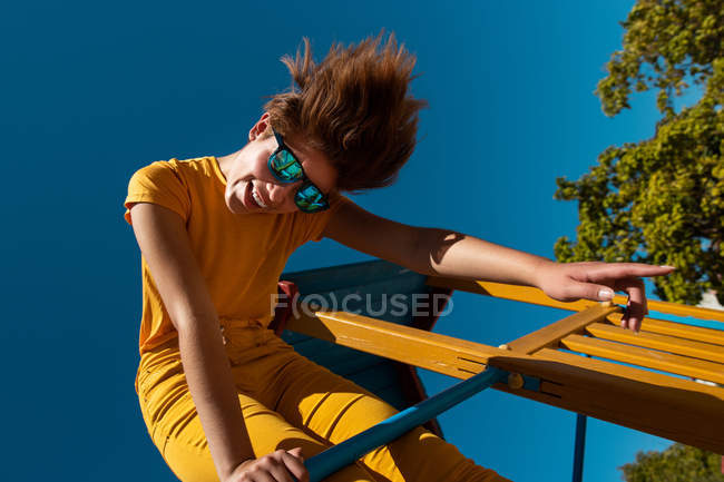From below trendy teenager in sunglasses playing on yellow crossbar against clear blue sky — Stock Photo