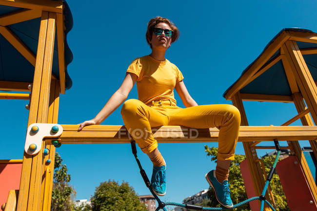 D'en bas adolescent branché dans des lunettes de soleil assis sur barre transversale jaune contre ciel bleu clair — Photo de stock