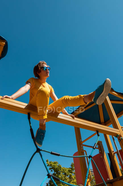 Снизу модный подросток в солнечных очках, сидящий на желтой перекладине на фоне ясного голубого неба — стоковое фото
