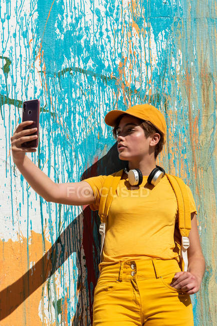 Стильный миллениал делать селфи со смартфоном, стоя напротив стены с красочными красками капает — стоковое фото