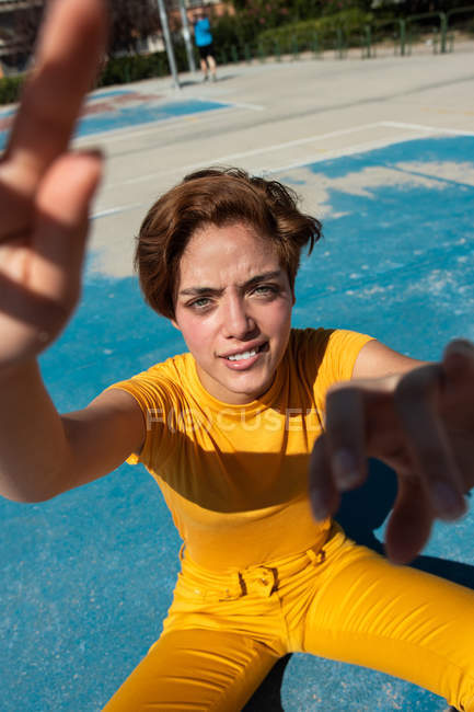 High angle of cool teenager in yellow clothes looking in camera and showing hands on blue sports ground — Stock Photo