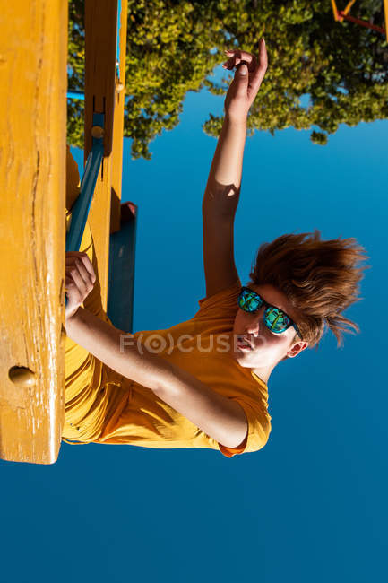 Desde abajo adolescente de moda en gafas de sol jugando en la barra transversal amarilla contra el cielo azul claro - foto de stock