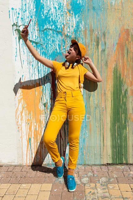 Elegante millennial tomar selfie con teléfono inteligente, mientras que de pie contra la pared con goteos de pintura de colores - foto de stock