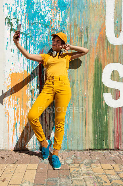 Stylischer Millennial macht Selfie mit Smartphone, während er mit bunten Farbtropfen an der Wand steht — Stockfoto