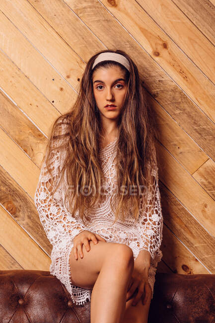 Jeune femme en robe blanche assise sur un canapé avec un fond en bois. — Photo de stock