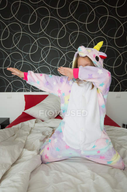 Fröhliches Mädchen im Einhorn-Pyjama, das Spaß hat und sein Gesicht auf dem Bett mit weißer und roter Bettwäsche bedeckt — Stockfoto